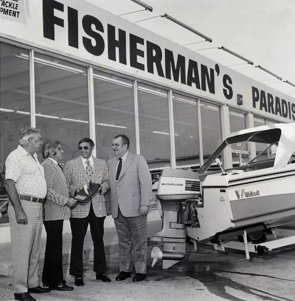 Four men in front of the Fisherman's Paradise storefront beside an Evinrude-powered Wellcraft